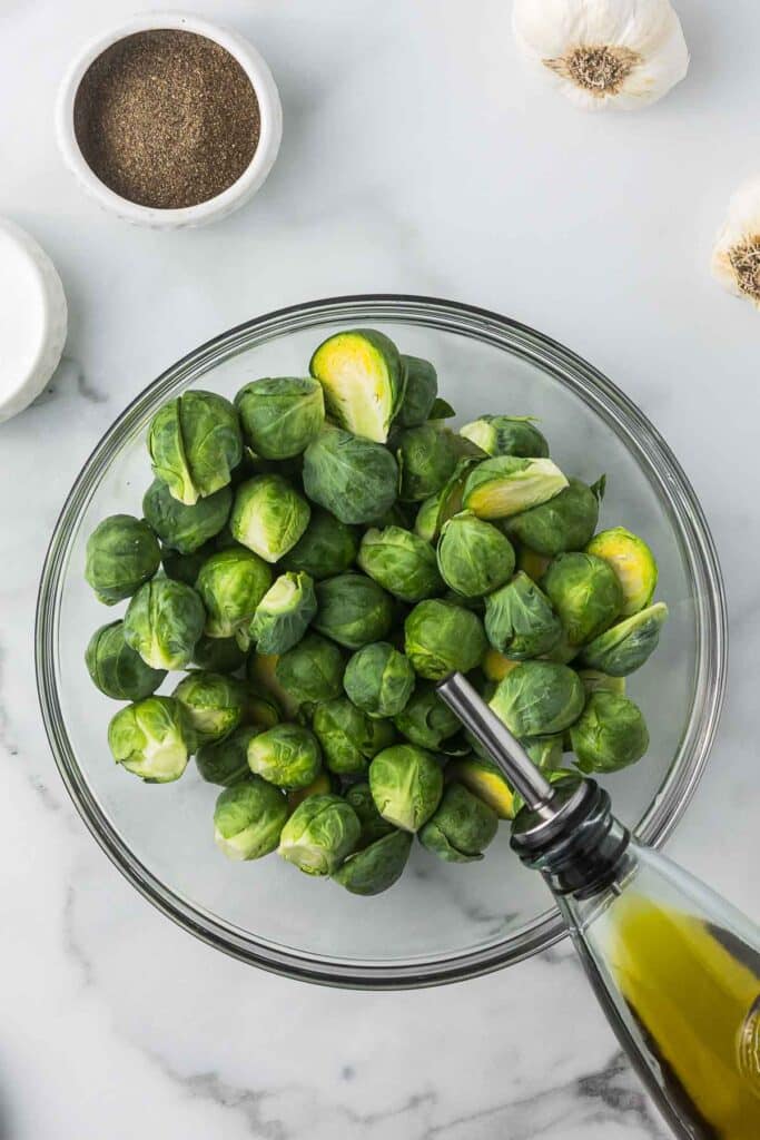 A clear bowl of Brussels spouts with olive oil being added.