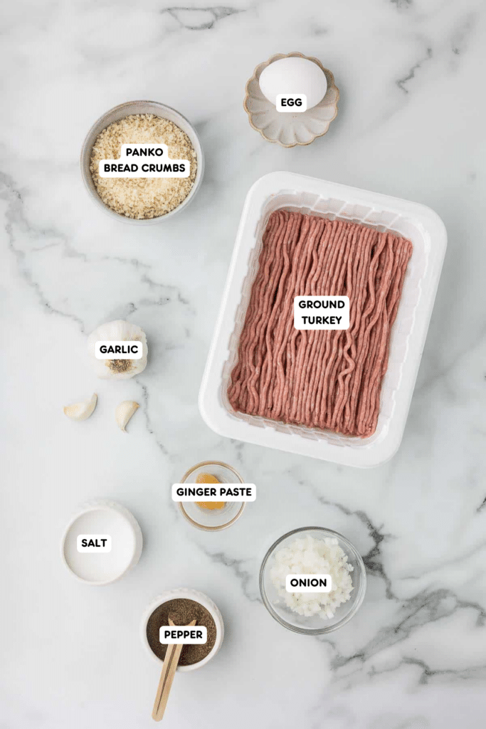 Ingredients for baked teriyaki turkey meatballs, including ground turkey, panko bread crumbs, egg, garlic cloves, ginger paste, diced onion, salt, and pepper, neatly arranged on a marble countertop.