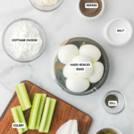 An image of ingredients for the recipe "Cottage Cheese Egg Salad with Dill Pickles:" all sat out on a marble counter. Ingredients are seperated, and listed with each item needed for the recipe from top left to bottom: mayonnaise, pepper, cottage cheese, hard boiled eggs, salt, celery, onion, dill, and relish. The celery and onion are on a brown cutting board.