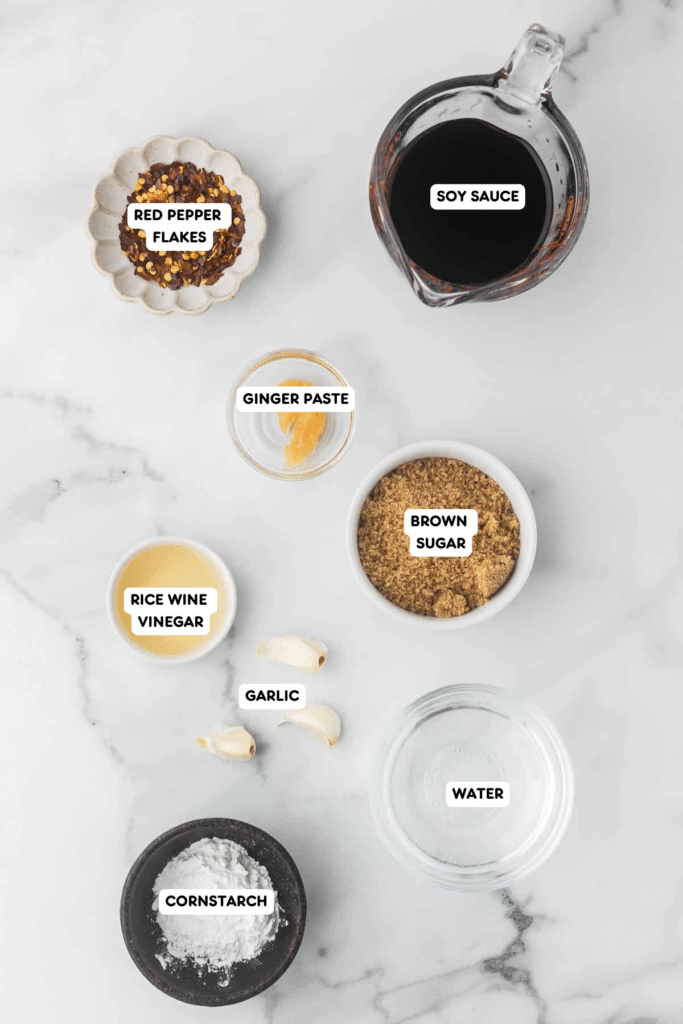 Ingredients for homemade teriyaki sauce, including soy sauce, brown sugar, rice wine vinegar, ginger paste, garlic cloves, red pepper flakes, water, and cornstarch, displayed on a marble countertop.