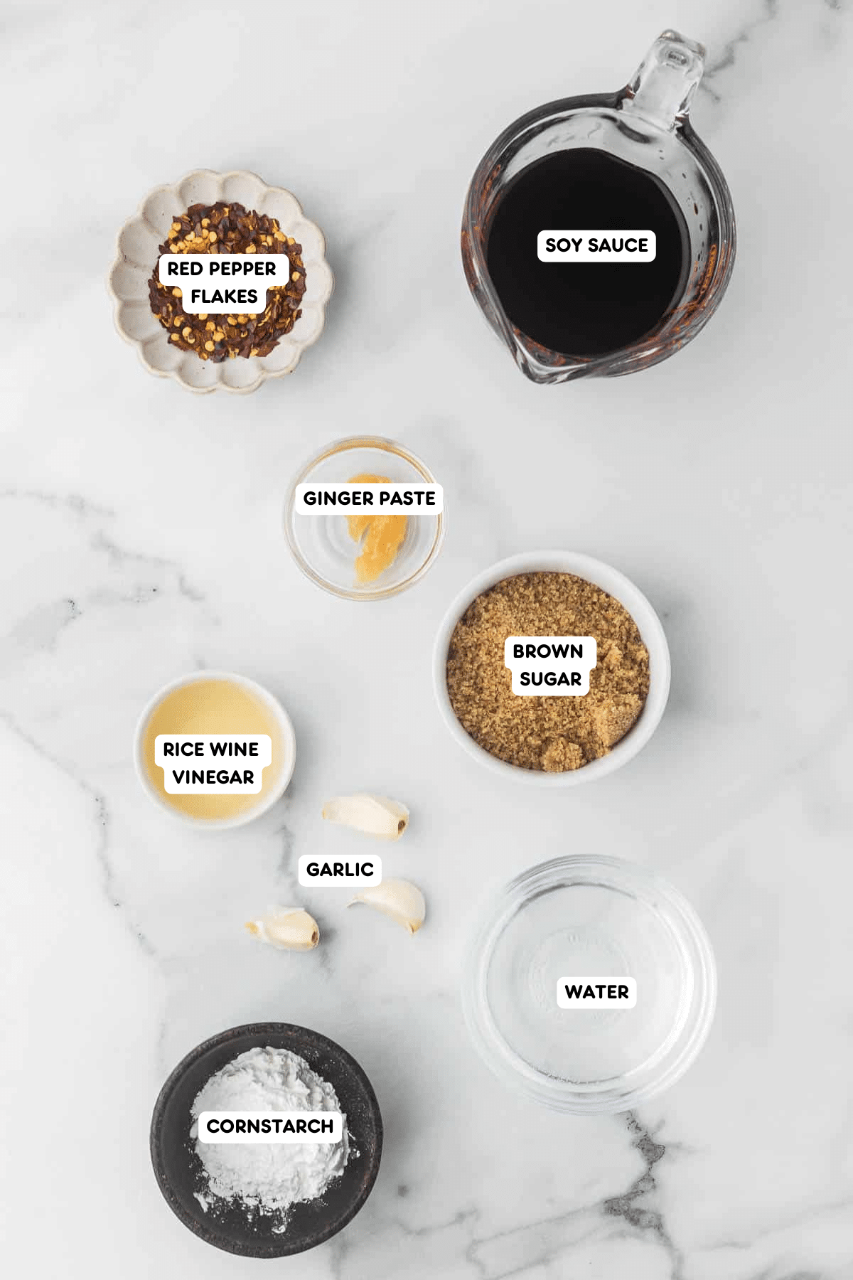 Ingredients for homemade teriyaki sauce, including soy sauce, brown sugar, rice wine vinegar, ginger paste, garlic cloves, red pepper flakes, water, and cornstarch, displayed on a marble countertop.