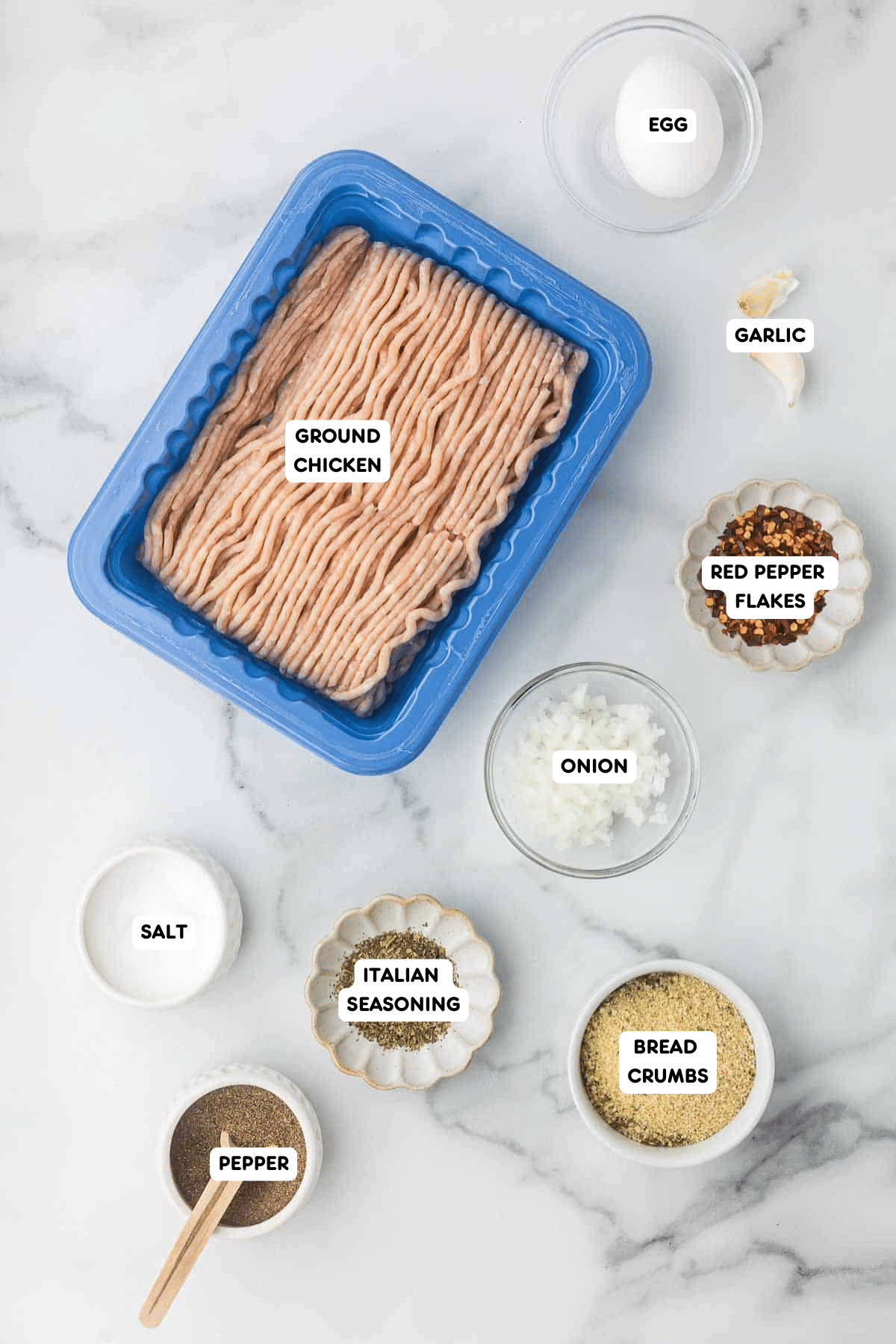 Ingredients for air fryer chicken meatballs, including ground chicken, bread crumbs, egg, garlic, red pepper flakes, onion, salt, black pepper, and Italian seasoning, arranged on a marble countertop.