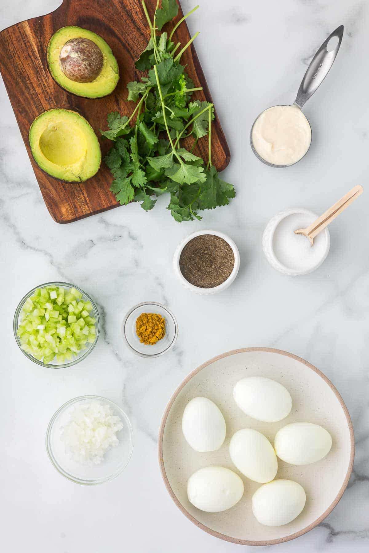 Ingredients to make egg salad including spices, eggs, onion, celery, avocado, mayo and cilantro.