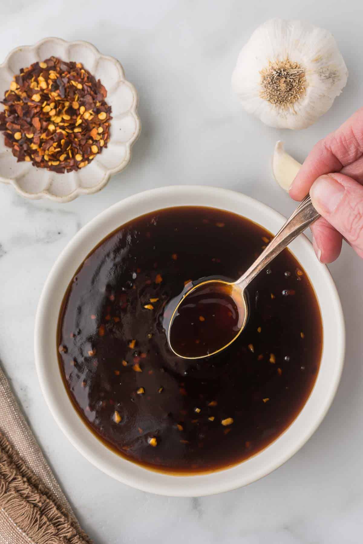 Homemade teriyaki sauce in a white bowl with a spoon and a bowl of red pepper flakes and garlic next to the sauce.