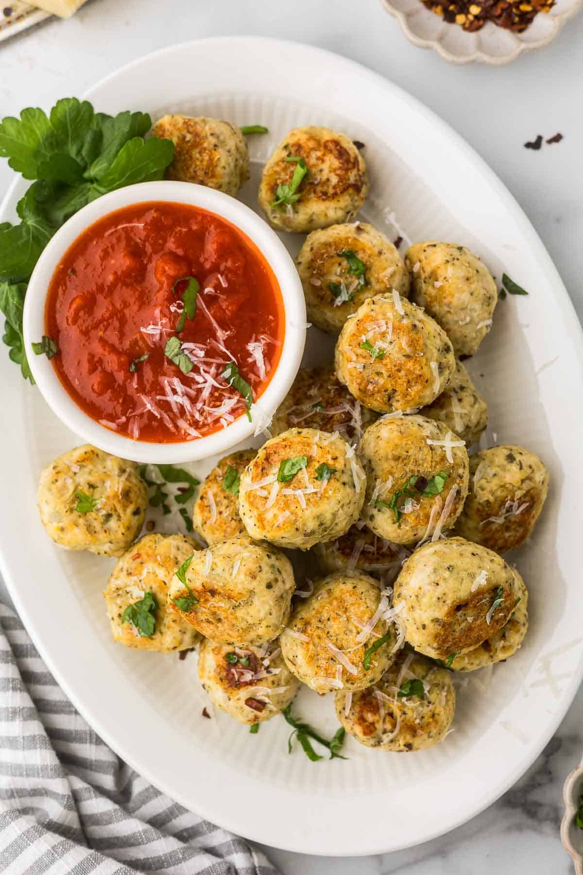 Baked Chicken Ricotta Meatballs on a white platter with homemade marinara sauce, fresh parsley and grated parmesan cheese.