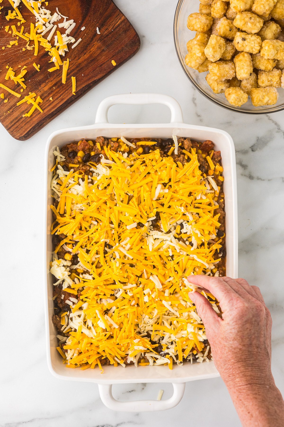 Cheese being added to the top of the ground beef mixture before adding tots.
