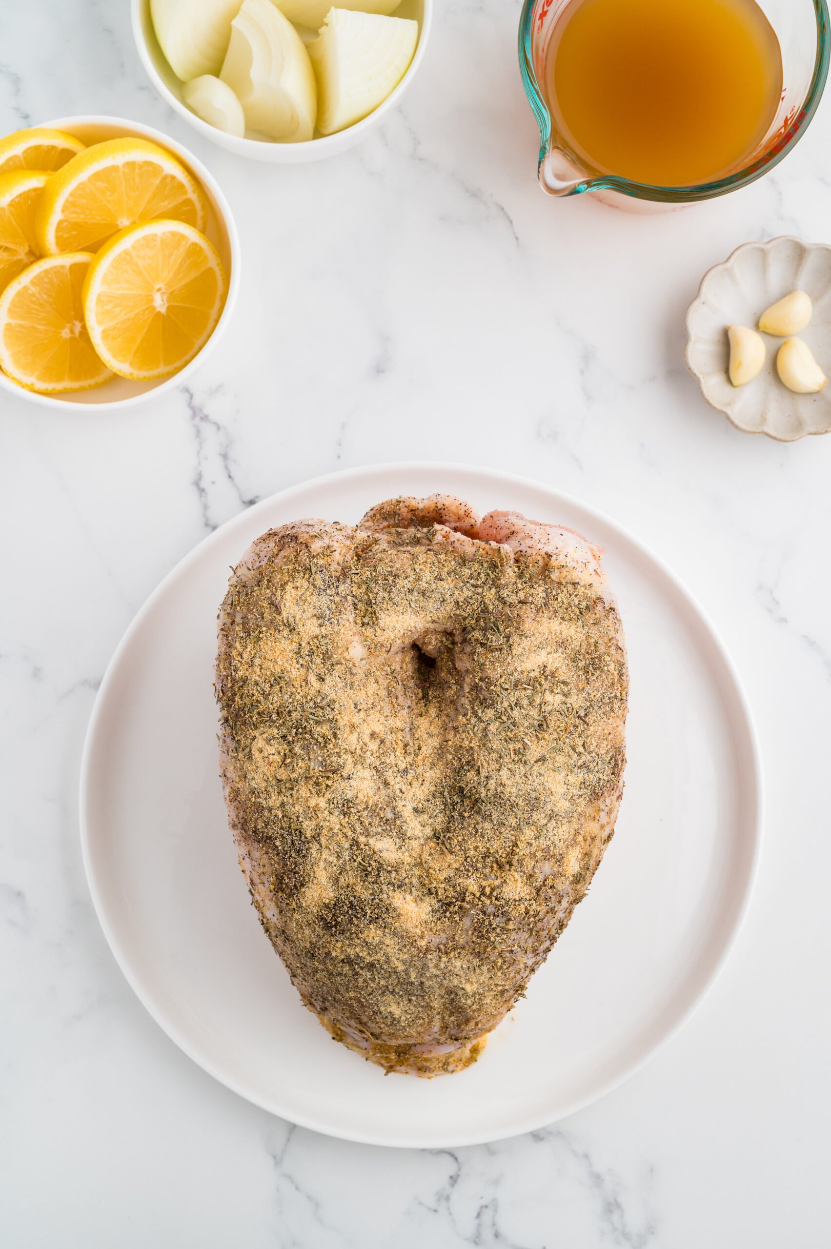 Bone-in turkey breast coated with herbs on a plate with lemon, onion, garlic, and broth.