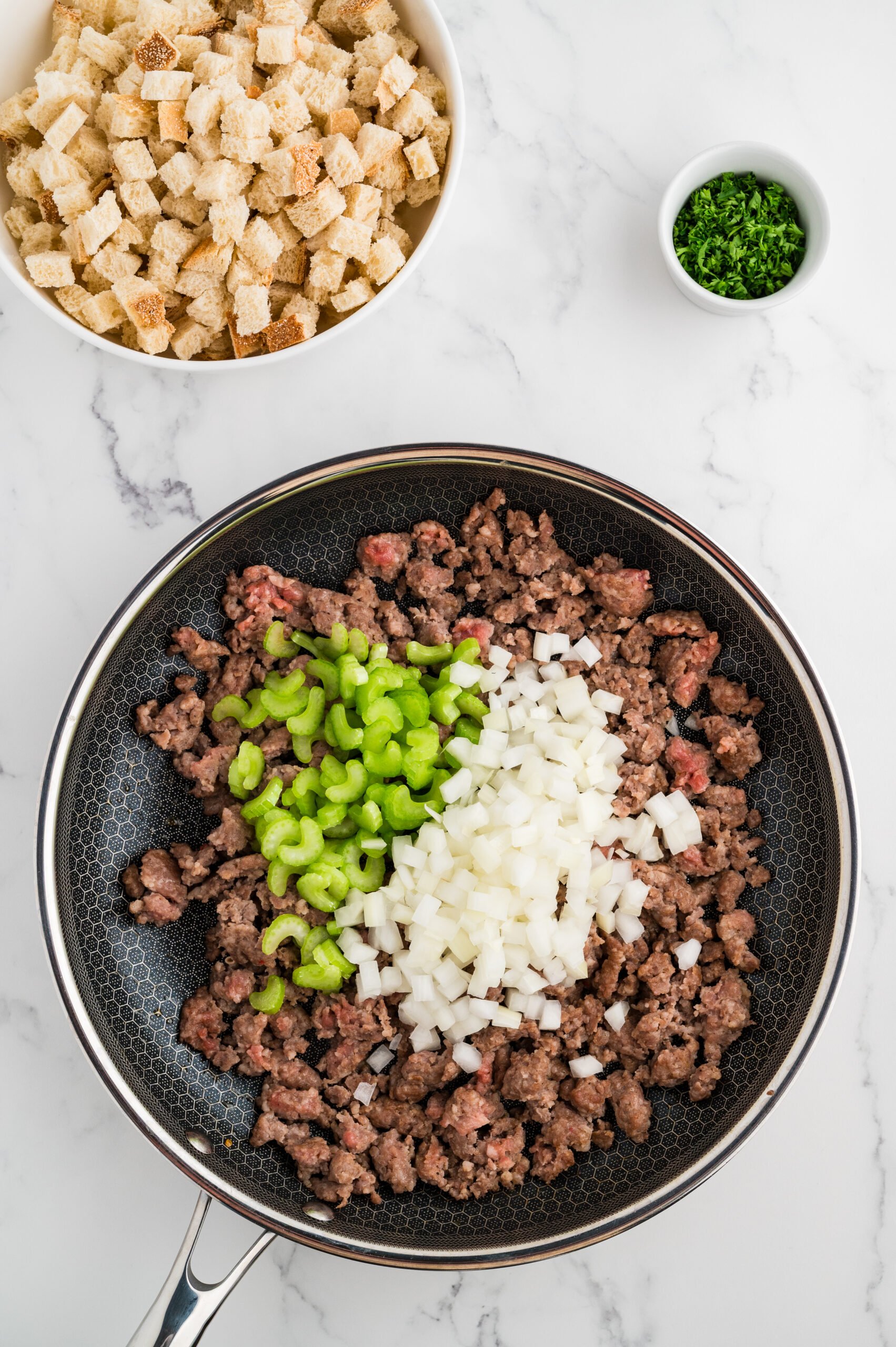 Ground sage pork sausage cooking in a skillet with chopped onion and sliced celery for stuffing.