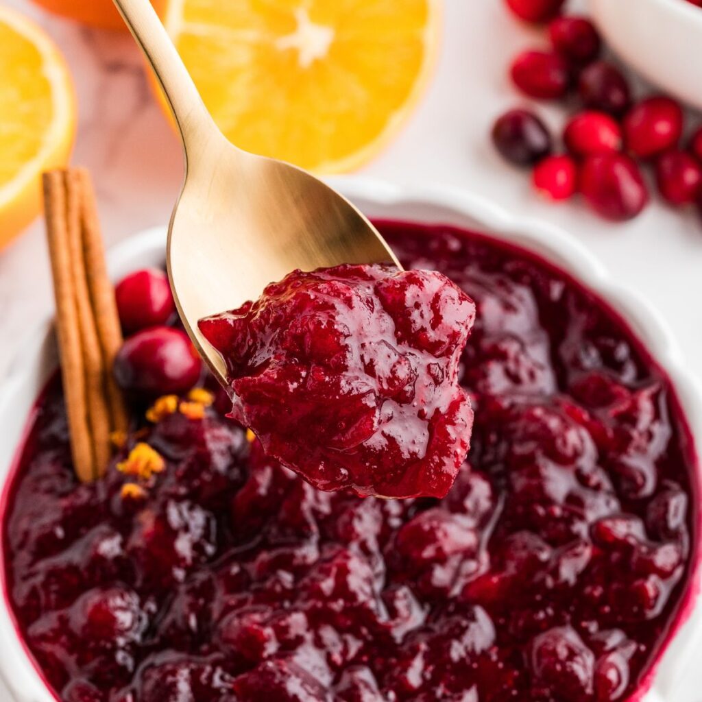 Gold spoon lifting a scoop of thick homemade whole berry cranberry sauce with oranges and cranberries in the background.