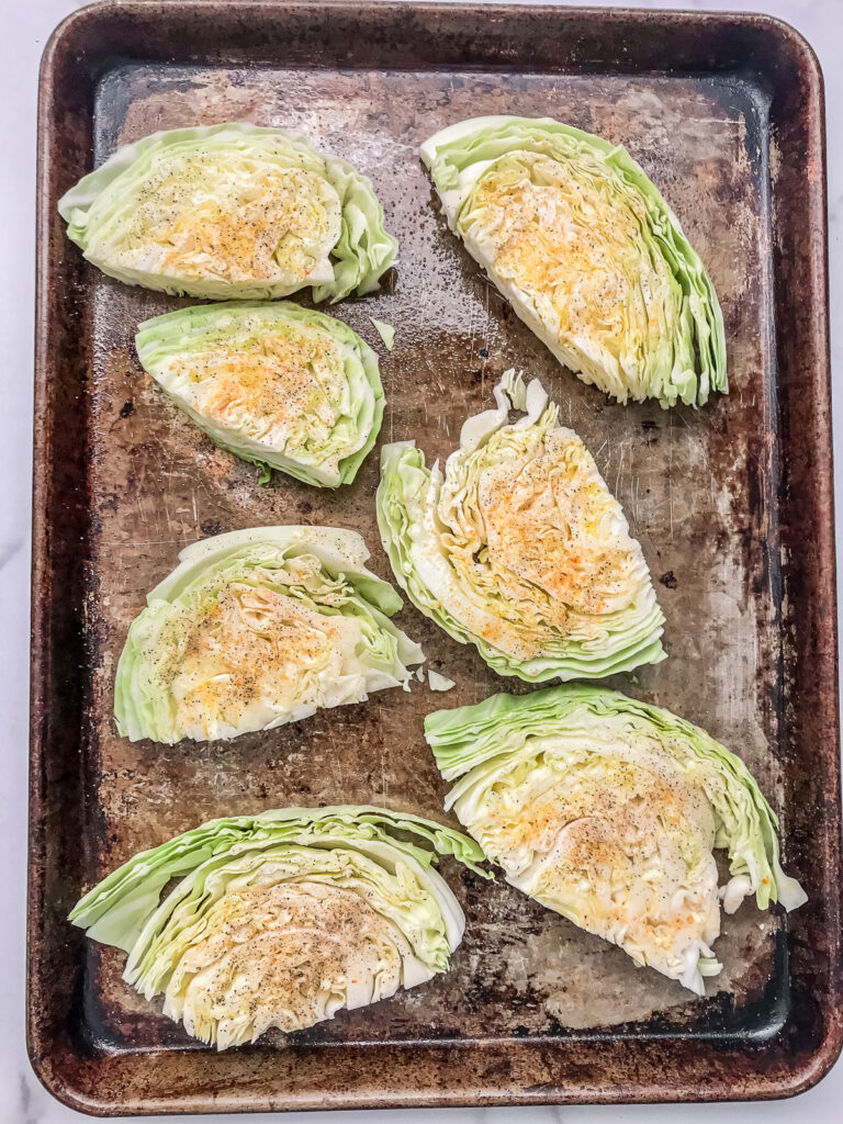 Cabbage steaks topped with bacon, olive oil, and seasoning on a baking sheet before roasting.