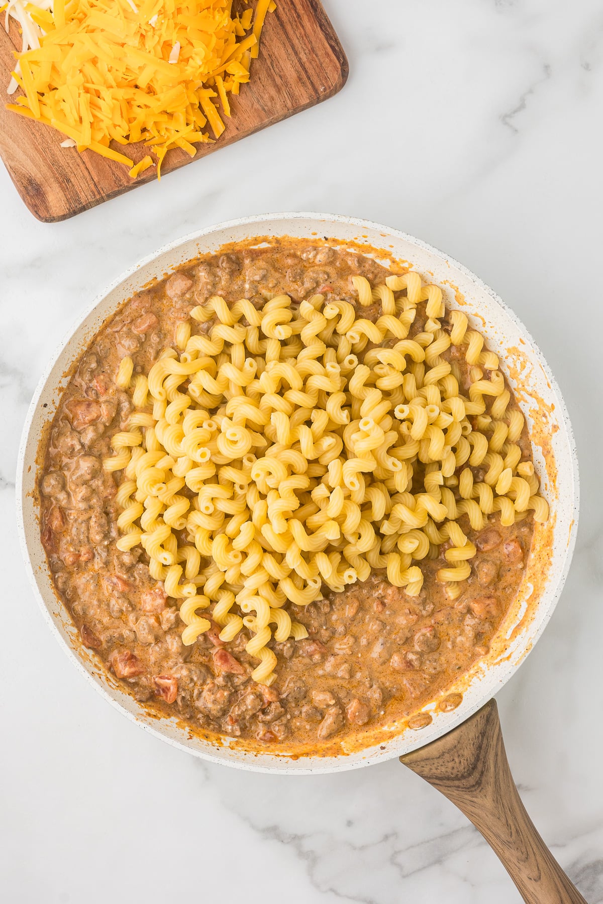 Cooked pasta added to creamy enchilada beef sauce in skillet for Mexican mac and cheese.
