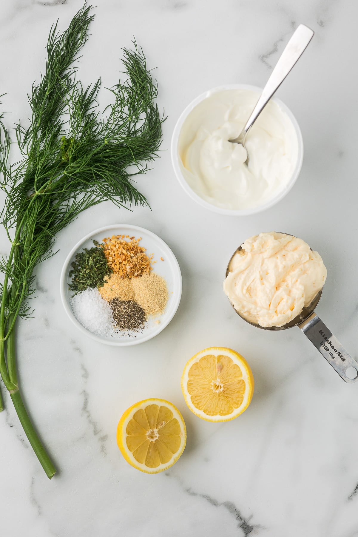 Mayonnaise, sour cream, spices, lemon and fresh dill on a table before creating a dill dip.