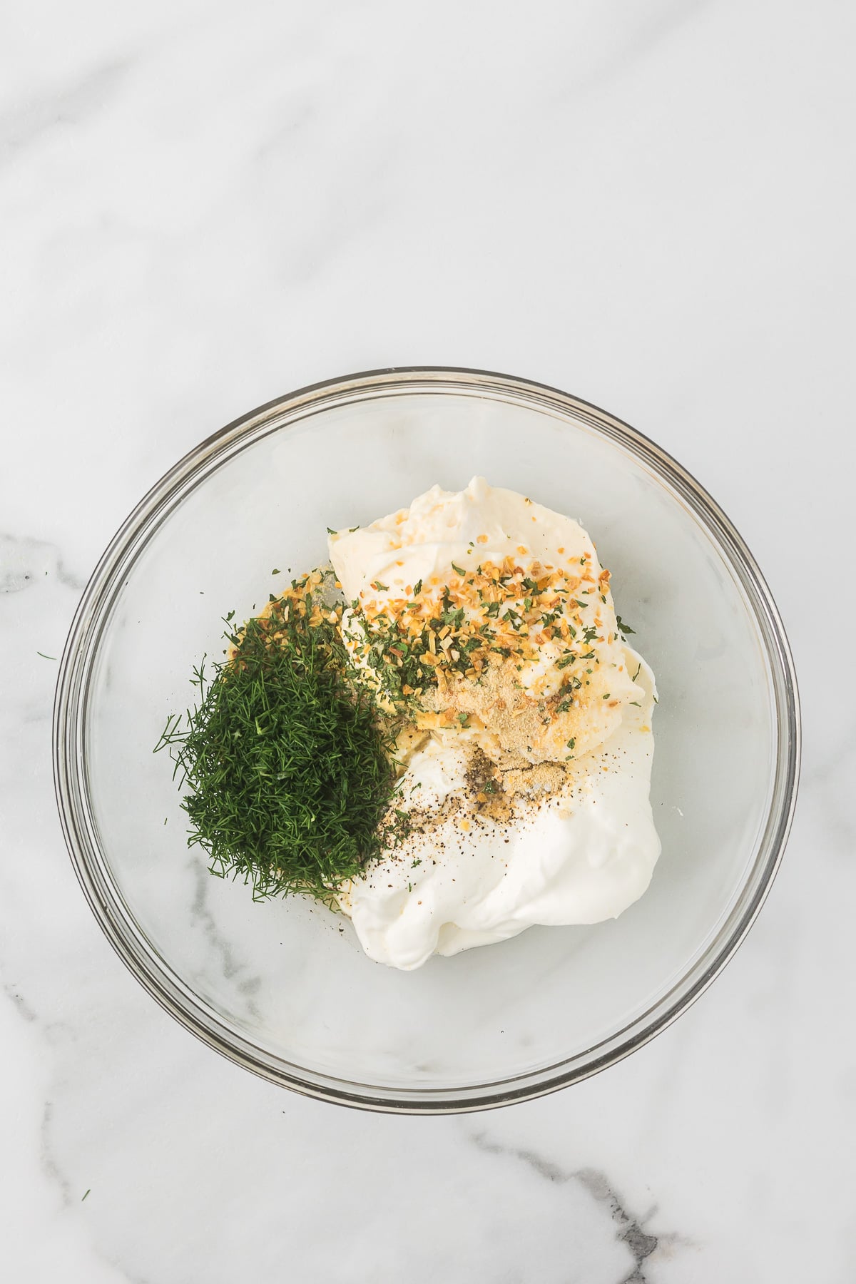 The ingredients in a glass bowl to make dill dip including mayonnaise, sour cream, spices, lemon juice and fresh dill.
