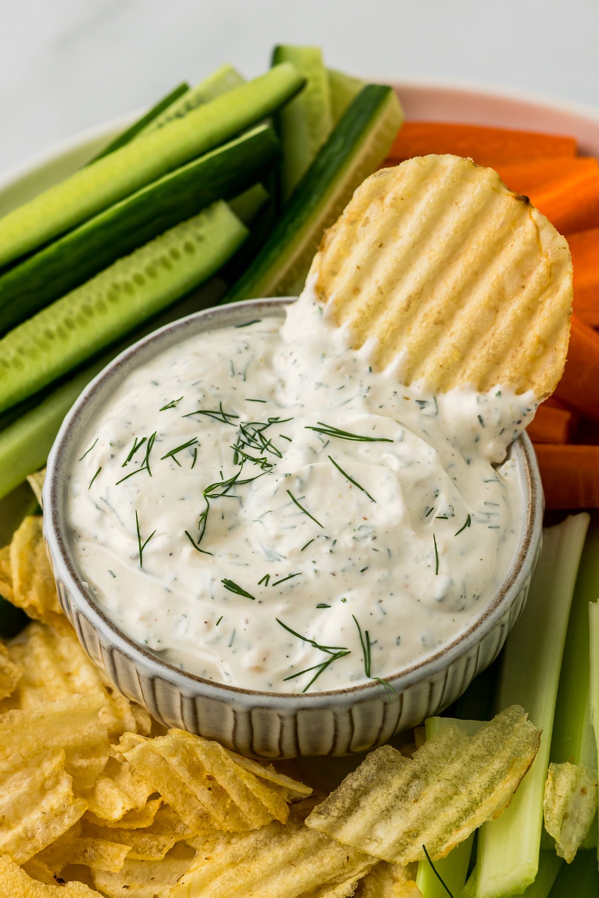 Dill dip in a bowl with a potato chip in the dip and veggies around the bowl.