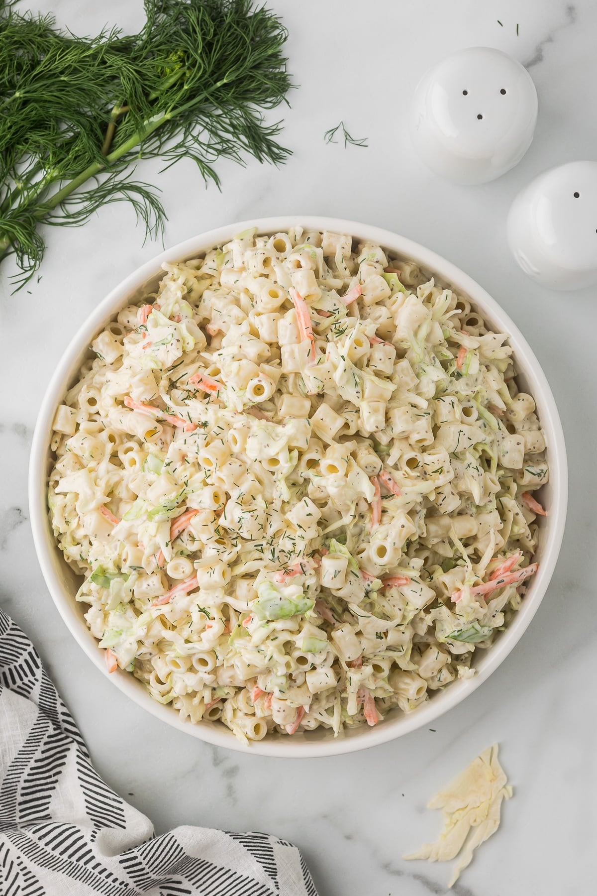 Creamy Colelsaw recipe with pasta in a white bowl with fresh dill and salt pepper shakers.