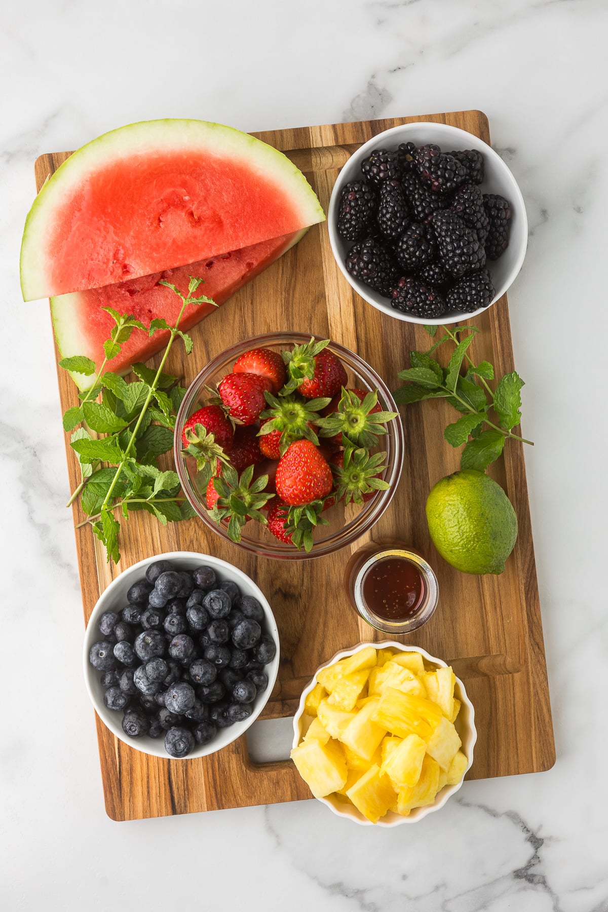 Fresh watermelon, strawberries, blueberries, blackberries, pineapple, mint, honey, and lime arranged on a wooden board for a summer fruit salad.