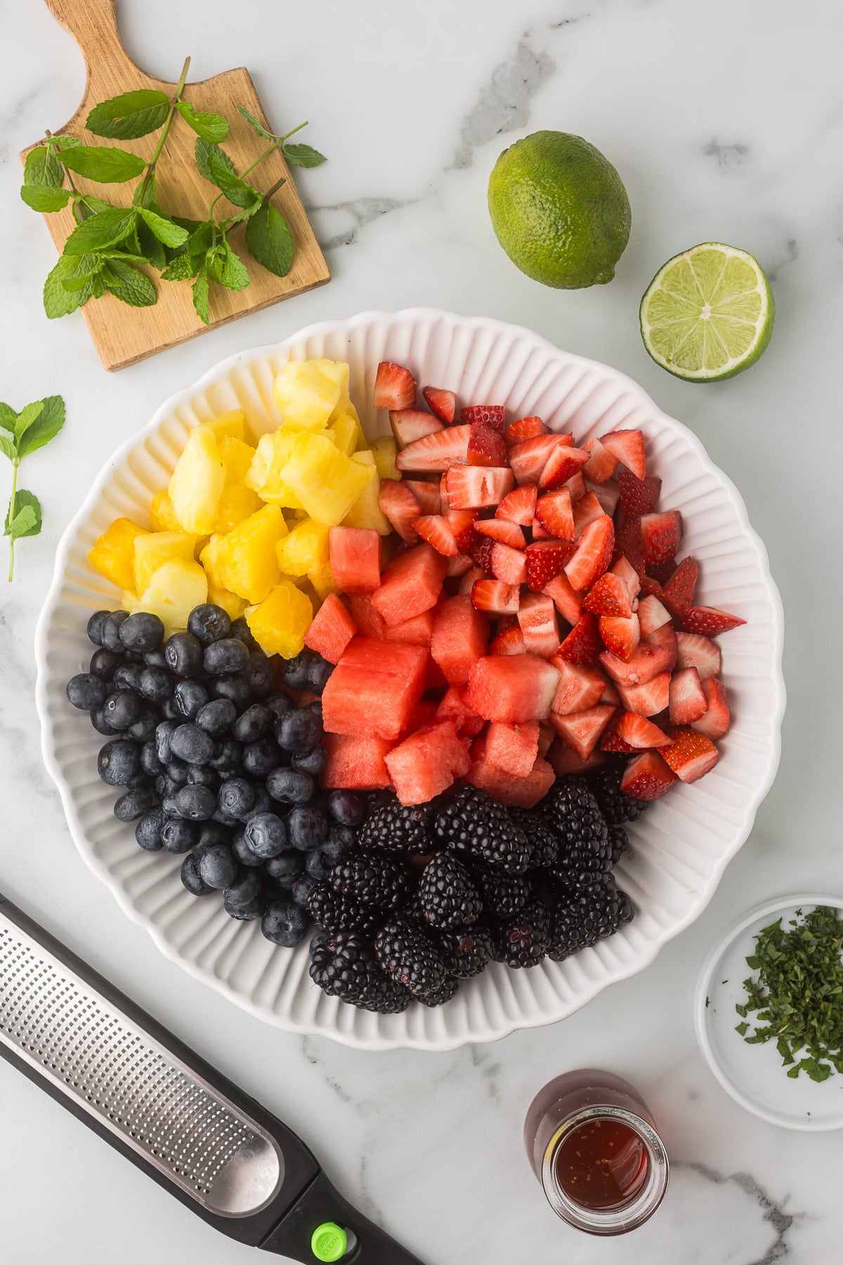 Ingredients for a berry fruit salad including watermelon, pineapple, strawberries, blueberries, and blackberries arranged in a serving bowl.