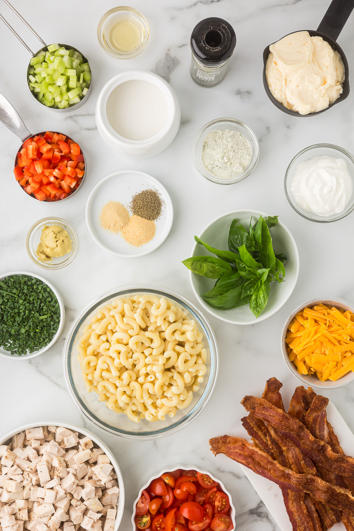 Ingredients for BLT Chicken Pasta Salad including cooked chicken, elbow macaroni, bacon, cheddar cheese, tomatoes, celery, red bell pepper, chives, basil, and creamy dressing ingredients.